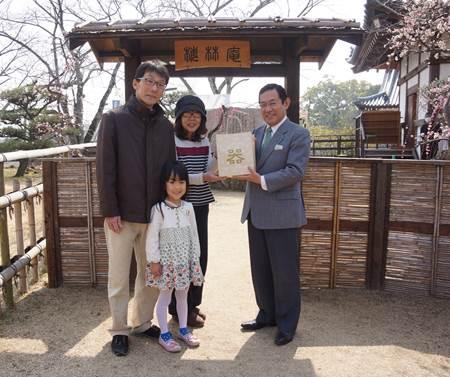 木と竹で作られた門の前で、スーツ姿の男性から記念品を受け取っている家族連れの写真