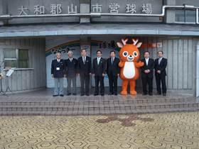 スーツを着た8人の実行委員とオレンジ色の鹿のゆるキャラの写真