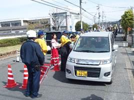 黄色い服を着た一団が、銀色の軽自動車のドライバーに交通安全のチラシを配っている処の写真