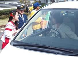 青い制服を着た警察官と、赤と白の衣装を着たキャンペーンガールが、ドライバーに交通安全のチラシを渡している様子