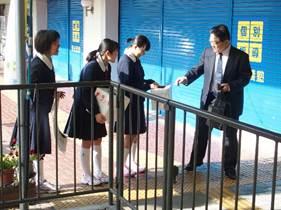 青色のシャッターが閉まった塾の前で、三人の女子生徒に募金するスーツ姿の男性の写真