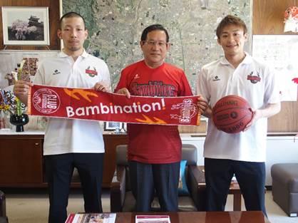 執務室で、バンビシャス奈良の赤いタオルとバスケットボールを持つ二人のバンビシャス奈良の選手と、その間に立つ赤いシャツを着た市長の記念写真