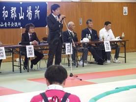 武道館で、マイクを持った黒いスーツを着た男性が開会の挨拶をしている処の写真