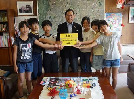 6人の小学生が市長が持った黄色い募金箱に手を添えている記念写真