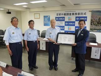 賞状を掲げる1人の青いスーツの男性と3人の制服の男性の写真