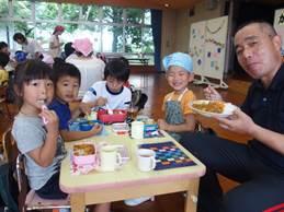 カレーを食べる4人の園児をバックに、カレーを食べる先生の写真
