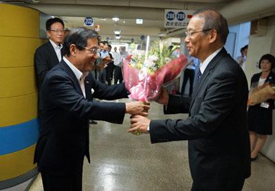 男性の市職員が同僚に見守られながら、黒いスーツの男性にピンクの花束を渡しているところの写真