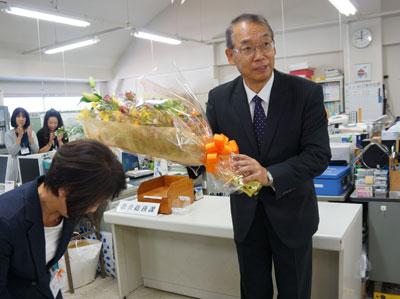 黒いスーツの男性が、女性の市職員から花束を贈られているところの写真