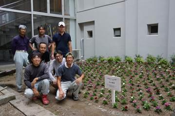 中庭にて植樹した花苗や低木の畑と若手農業者グループ7人が記念撮影をしている様子の写真
