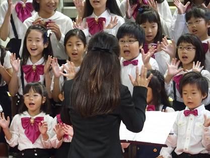 完成を祝って、黒い服の女性が指揮をし、三の丸少年少女合唱団の子供たちが合唱を披露している写真