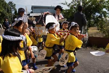 黄色い法被を着た園児たちが手作りしたおみこしを担いで豊作を祝っている写真