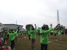 運動場で参加者や関係者を含む人たちがラジオ体操をしている写真