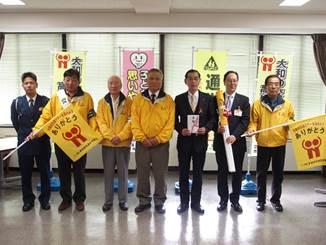 横断旗贈呈式で黄色い上着を着た男性4人と市長や警官が並んでいる写真