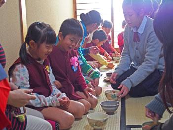 片桐中学校の和室で茶道体験し、並んで正座をしている園児たちと抹茶を振る舞う茶道部の女子生徒の写真
