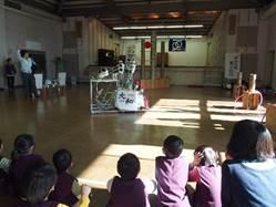 屋内ホールで 奈良高専の生徒が作ったロボットが輪投げを披露しているところと見入っている園児たちの写真