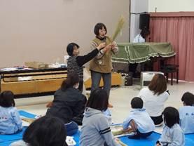 幼稚園の屋内で藁を持ち「しめ縄作り」を説明している女性職員2人とその工程を見ている園児たちの写真