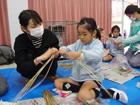 女性職員に教わりながら女児と一緒にしめ縄を編んでいる写真