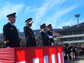 紅白の垂れ幕が取り付けられた高台に4名の代表者が敬礼して式典を観閲する様子の写真