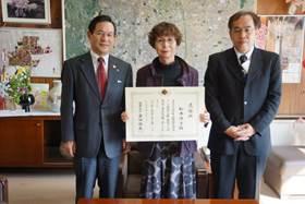 紫のコートを着て、感謝状を見せる松本さんを中心に、市長と人権擁護課山本課長の記念写真