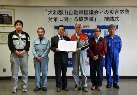 「大和郡山自動車協議会との災害応急対策に関する協定書」締結式と書かれた垂れ幕の前で協定書を見せている6人の関係者の記念写真