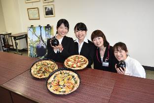 机の上に置かれたナスを使用した3枚のピザと、大和丸なすをに持ってこちらに笑顔を向ける4人の女子大学生の写真