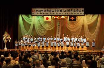 水色の制服を着た幼い児童たちがステージ上で一列に並んで合唱をしている様子と、それを見ている観客の写真