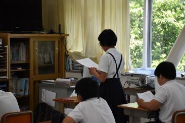 窓から緑が見える教室で、女子小学生が立ち上がって自作の詩を朗読している様子の写真