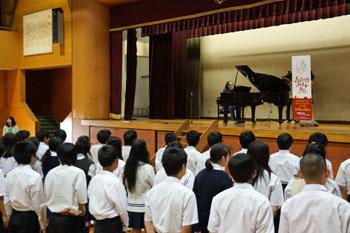 ステージの上にピアノがあり、演奏に合わせて合唱をしている中学生たちを背後から撮影した写真
