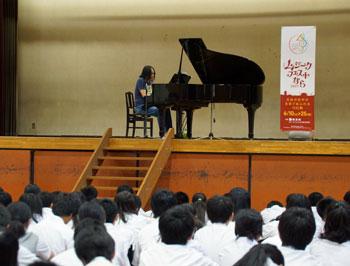 体育館の舞台上でピアノを演奏している男性と、その演奏を体育座りで聞いている中学生たちの様子の写真