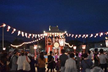 中央に太鼓が置かれた一段高い櫓が立ち、会場内に渡された提灯が灯っている中で盆踊りする人々の写真