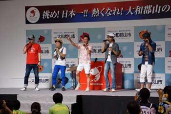 赤色のTシャツや青いパンツを履いた男性5人が舞台上で歌っている写真