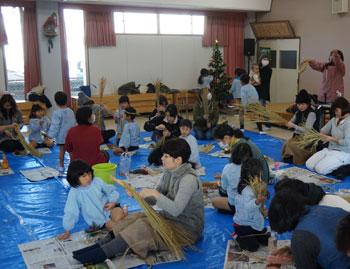 ブルーシートが敷かれた床に座っている大勢の親子連れがしめ縄を作っている写真