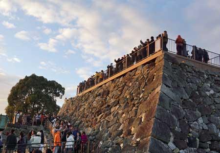 雲がかかった空の下柵のある城の土台の上に集まる大勢の男女と土台に続く階段に並ぶ大勢の男女の写真