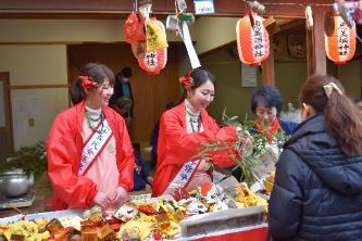出店で祭りの来場者の女性の対応をする紅白のたすきの上から赤いジャンパーを羽織る2人の女性の写真