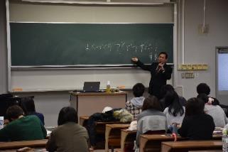 背後の黒板に書き込まれた文字に右手を近づけ左手でマイクを持ったスーツ姿の男性と教室の机に座る男女の学生たちの写真