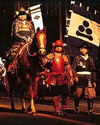 後ろに幟が立っていて、鎧や兜をつけた人が馬に乗っていて、右に鎧や兜をつけた人が2人いる写真