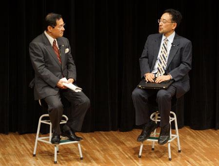 舞台上の黒いカーテンを背景に小さな椅子に座って見つめ合う2人のスーツ姿の男性の写真