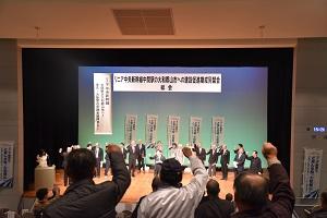 リニア中央新幹線中間駅の大和郡山市への建設促進期成同盟会・講演会・総会 と書かれた看板の下、同じ内容ののぼりを背にして拳を掲げる講演者と椅子から立ち上がる聴者たちの写真