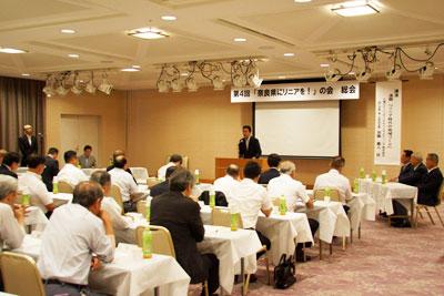 暖色を基調とした部屋の中、奈良県にリニアを！の会と書かれた横看板とプロジェクターを前に説明する講演者と聴者たちの写真