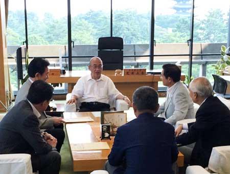 大きな窓から外の景色が見える部屋で、机を囲って話し合いをする6人の男性の写真