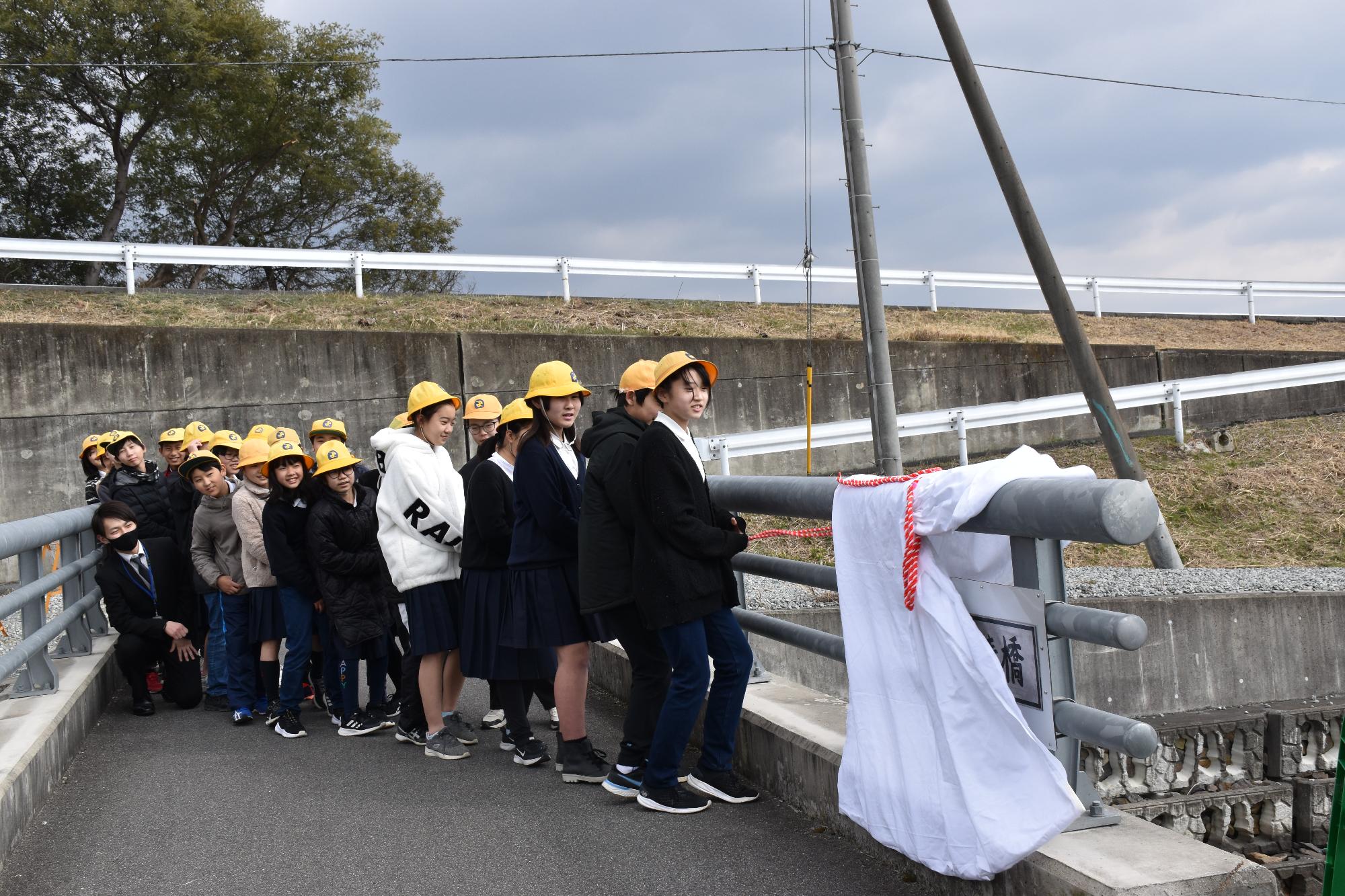 橋名板にかけられた白い布をロープを引っ張り除幕する小学生25人