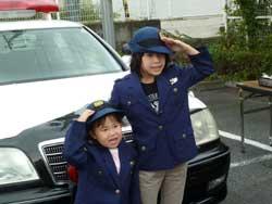 パトカーの前で婦人警官の制服を着て敬礼をしている2人の子供たちの写真