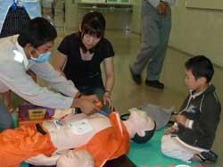 「救急講習」で心肺蘇生法を教えている講師役の救急隊員とそれに聞き入っている保護者と子ども写真