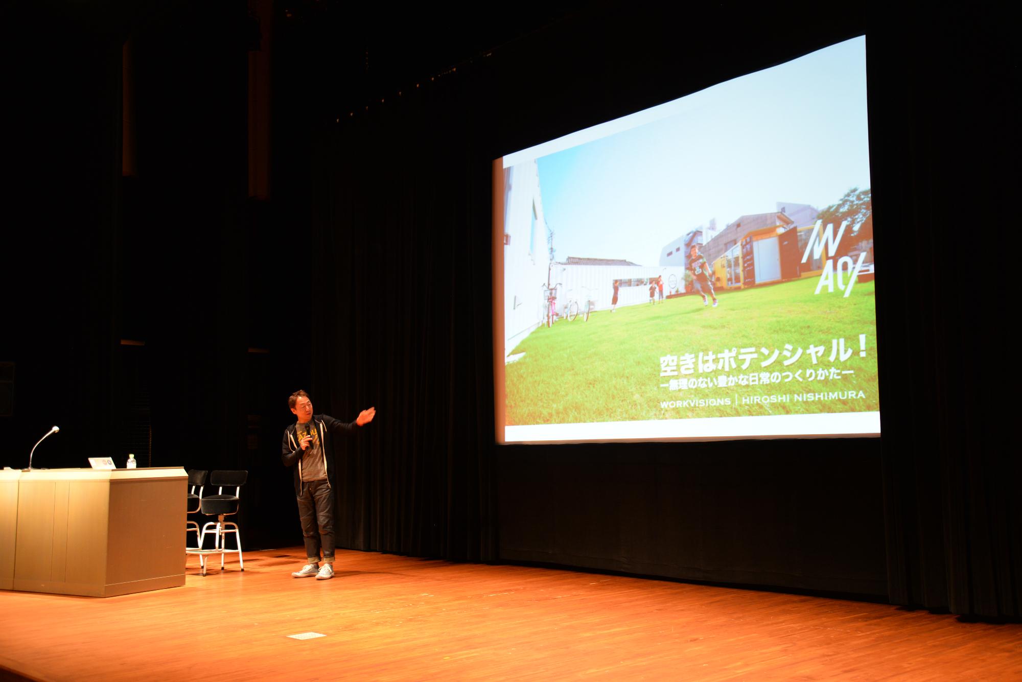 ステージ上で講師の西村浩氏がスクリーンに向かって手を向けている。