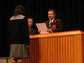 舞台上で表彰状を読み上げるスーツ姿の男性と、受賞者の女の子の写真