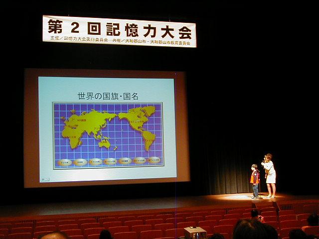 舞台上のスクリーンに映し出された世界地図と、暗唱発表をしている男の子の写真