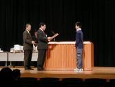 舞台上で表彰状を読み上げる男性、トロフィーを持った男性と受賞者の男の子の写真