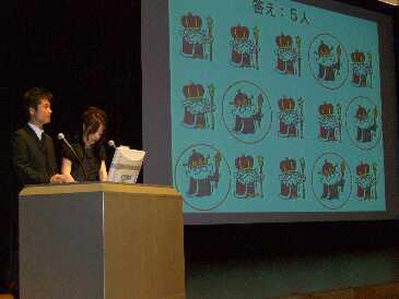 キャラクターのイラストが映し出されたスクリーンと、その横で書類を読む司会者2人の写真