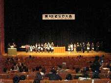 舞台上で表彰状を手に横に並んで立つたくさんの入賞者の人々を遠くから撮影した写真