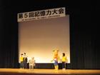 舞台上に立つ、数人の人とキャラクターの着ぐるみをきた人物の写真